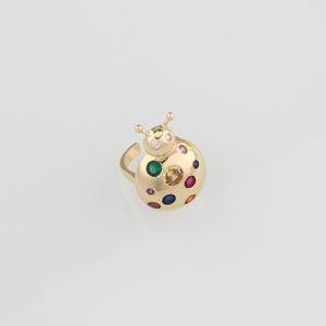 May include: A gold-colored ring shaped like a ladybug. The ring features a round body with colorful gemstones and two small antennae. The gemstones are in various colors, including red, green, blue, and yellow.