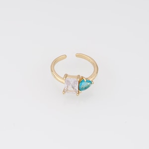 Puede incluir: Un anillo abierto de tono dorado que presenta dos piedras distintas: una gema transparente de corte rectangular y una gema más pequeña de color turquesa con forma de pera. La banda es fina y metálica, con una abertura ajustable en la parte superior.
