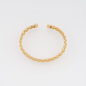 May include: A gold-colored cuff bracelet with a braided design. The open-ended bracelet has a rounded shape and is made of interwoven strands. The ends of the bracelet have small, rounded caps.