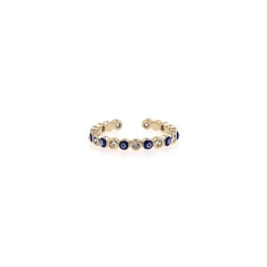 May include: A gold-colored open ring featuring a series of alternating blue evil eye and clear crystal accents. The ring is set against a white background, highlighting its delicate design and intricate details. A stylish piece of jewelry.
