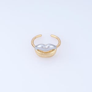 May include: A gold-toned open ring featuring a central design of lips. The upper lip is finished in a smooth silver tone, while the lower lip is textured with vertical lines and finished in a polished gold tone, matching the thin circular band.