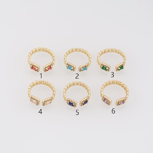 Può includere: Sei anelli aperti color oro con un design a fascia a catena intrecciata. Ogni anello presenta due pietre preziose rettangolari taglio baguette alle estremità, numerate da 1 a 6. Le pietre sono rosse, turchesi, verdi, trasparenti, blu scuro e viola.