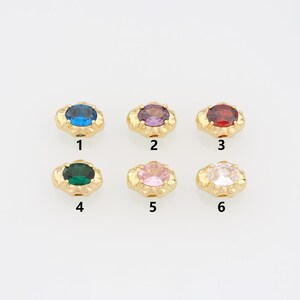 May include: Six gold-toned metal charms, each featuring a central oval-cut gemstone in blue, purple, red, green, pink, or clear. Each charm is numbered 1 through 6, with a textured, irregular gold border surrounding the faceted stone center.