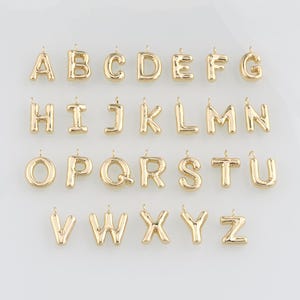 Könnte beinhalten: Eine Sammlung goldfarbener Buchstabenanhänger, jeweils mit einer Schlaufe zum Befestigen an einer Halskette oder einem Armband. Die Buchstaben sind in einer abgerundeten, ballonartigen Schriftart angeordnet und in Reihen von A bis Z angeordnet.