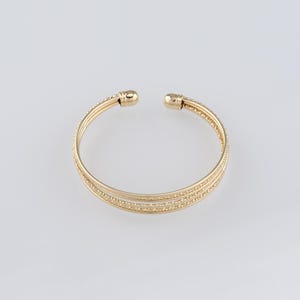 Op de afbeelding: Een gouden armband met een getextureerd ontwerp. De armband is gemaakt van meerdere dunne banden die met elkaar zijn verweven.