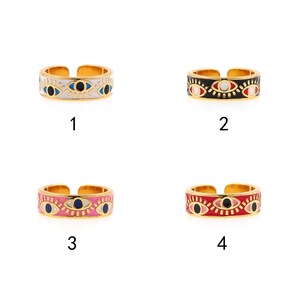 May include: Four gold-toned adjustable rings with enamel eye designs. Each ring features a different color scheme: white, black, pink, and red. The eye designs include blue, black, and white details. The rings are displayed against a white background.