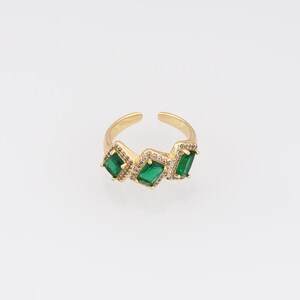 May include: A gold-toned open ring featuring three emerald-cut green stones, each surrounded by a halo of small clear crystals. The band is adjustable with a gap at the top, showcasing a polished metallic finish and a symmetrical arrangement.
