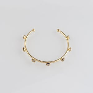Puede incluir: Un brazalete dorado con pequeños detalles de piedras preciosas azul oscuro. El brazalete tiene un diseño sencillo y abierto con extremos redondeados. Las piedras preciosas están espaciadas uniformemente.