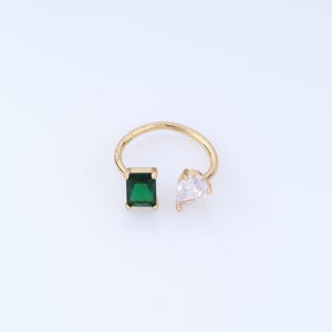 Può includere: Un anello aperto in oro con due pietre distinte alle estremità. Un lato presenta una pietra rettangolare verde smeraldo, mentre l'altro lato mostra una pietra trasparente a forma di goccia. La fascia è sottile e lucida.