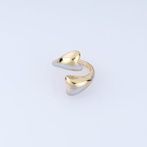May include: A two-tone open ring featuring a polished gold-colored top surface and a silver-colored bottom surface. The ring has a bypass design with two rounded, heart-like ends that curve toward each other, creating a smooth, reflective finish.
