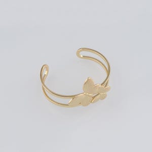 May include: Gold-coloured cuff bracelet with a butterfly design. The open-ended bracelet has two parallel bands and a decorative butterfly element with detailed wing patterns. The jewellery is set against a white background.