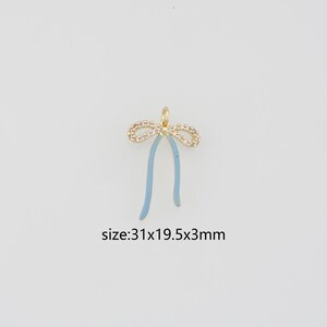 May include: A gold-toned metal charm in the shape of a bow, featuring clear cubic zirconia stones along the top loops and light blue enamel on the two hanging tails. The charm includes a small jump ring at the top and measures 31x19.5x3mm.
