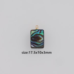Puede incluir: Un colgante rectangular con incrustaciones de concha de abulón iridiscente, que mide 1,75 cm x 1 cm x 0,3 cm, y un lazo dorado en la parte superior. La concha muestra tonos de azul, verde y marrón.