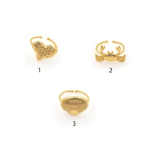 May include: Three gold-toned rings with different designs: a heart, a crab, and a seashell. The rings are displayed on a white background.