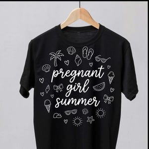 Peut inclure: T-shirt noir avec l'inscription blanche "pregnant girl summer". Le motif comprend des illustrations estivales telles que des coquillages, des lunettes de soleil et des cornets de glace. Le t-shirt est suspendu à un cintre en bois.