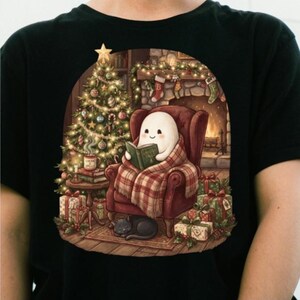 Peut inclure: Un t-shirt noir avec une illustration représentant un fantôme blanc assis dans un fauteuil rouge, enveloppé dans une couverture à carreaux, lisant un livre. Le décor inclut un sapin de Noël, une cheminée avec des chaussettes, des cadeaux et un chat noir.