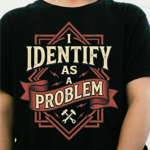 Puede incluir: Una camiseta negra de manga corta con un diseño gráfico que incluye el texto I IDENTIFY AS A PROBLEM en crema y rojo. El diseño presenta un borde en forma de diamante, una cinta decorativa y dos martillos cruzados en la parte inferior.