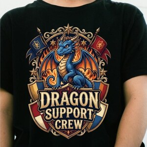 Peut inclure: Un t-shirt noir arborant un motif de dragon bleu centré dans un blason orné. Le design comprend deux drapeaux, des flammes et le texte DRAGON SUPPORT CREW dans une police serif grasse. L'illustration est détaillée avec des accents dorés et rouges.