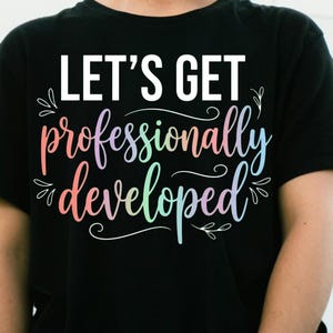 Peut inclure: Un t-shirt noir à manches courtes avec le texte blanc "LET'S GET" au-dessus de l'inscription cursive multicolore "professionally developed" dans des tons rose, violet, vert et bleu, agrémenté de petites fioritures décoratives blanches.