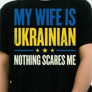 Peut inclure: Un t-shirt noir avec le texte MY WIFE IS UKRAINIAN en lettres bleues et jaunes à l'aspect vieilli, suivi de trois étoiles jaunes et du texte NOTHING SCARES ME en blanc, le tout centré sur le devant du vêtement.