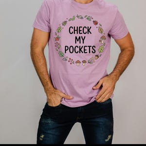 Peut inclure: Un t-shirt lilas avec le texte "CHECK MY POCKETS" en noir, entouré d'une couronne de feuilles, de baies et d'insectes. La personne porte un jean bleu foncé.