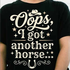 Peut inclure: Un t-shirt noir à manches courtes avec un texte couleur crème indiquant "Oops, I got another horse..." accompagné d'un graphique représentant un chapeau de cowboy, de petites étoiles et un fer à cheval, le tout dans une police décorative.