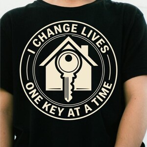 Peut inclure: Un t-shirt noir avec un motif circulaire crème. Le design représente la silhouette d'une maison avec une clé au centre. Le texte I CHANGE LIVES est inscrit en haut et ONE KEY AT A TIME en bas, à l'intérieur de la bordure circulaire.