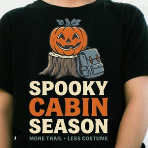 Peut inclure: Un t-shirt noir avec un motif représentant une citrouille d'Halloween orange posée sur une souche d'arbre brune à côté d'un sac à dos gris. Le texte "SPOOKY CABIN SEASON" est écrit en grandes lettres, suivi de "MORE TRAIL • LESS COSTUME" en plus petit.