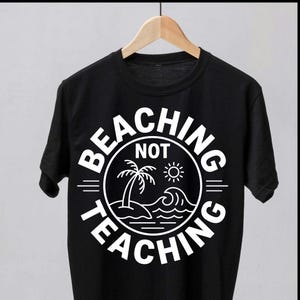 Peut inclure: T-shirt noir avec un motif blanc circulaire portant l'inscription "BEACHING NOT TEACHING". Le motif comprend un palmier, le soleil et des vagues, évoquant un thème de plage. Le t-shirt est suspendu à un cintre en bois.