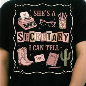 Peut inclure: Un t-shirt noir avec une impression graphique portant le texte "SHE'S A SECRETARY I CAN TELL". Le motif comprend une machine à écrire rose, des lunettes œil-de-chat, un pot à crayons floral, des bottes de cow-boy roses, une enveloppe, un carnet "Plans" et un cactus.