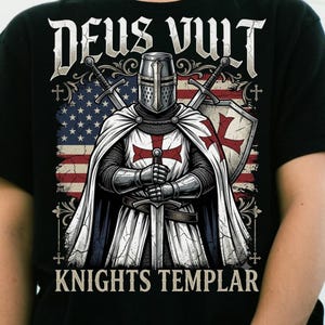 May include: A black t-shirt featuring a graphic of a knight in armor holding a sword, wearing a white cloak with a red cross. Behind the knight is an American flag and two crossed swords. The text DEUS VULT and KNIGHTS TEMPLAR is printed.