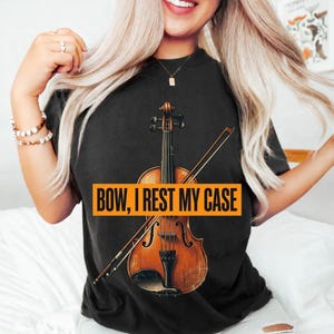 Könnte beinhalten: Ein schwarzes T-Shirt mit einer Grafik einer braunen Holzgeige und einem diagonal darüber liegenden Bogen. Über der Mitte der Geige befindet sich ein rechteckiger orangefarbener Block mit dem schwarzen Text: BOW, I REST MY CASE.