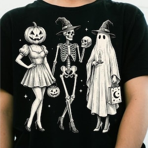 Könnte beinhalten: Ein schwarzes T-Shirt mit einer weißen grafischen Illustration von drei Figuren: eine Person mit Kürbiskopf in einem Kleid, ein Skelett mit Hexenhut und Absätzen, das einen Schädel hält, und ein Geist mit Hexenhut, der eine Kerze und eine Tasche hält.