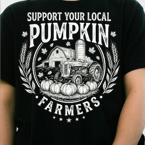 Peut inclure: Un t-shirt noir avec une impression graphique blanche représentant une scène de ferme avec un tracteur, une grange, un silo et des citrouilles. Le texte indique SUPPORT YOUR LOCAL PUMPKIN FARMERS. Le motif est entouré d'épis de blé et de feuilles.
