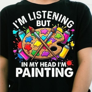 Könnte beinhalten: Ein schwarzes T-Shirt mit einer Grafik aus einer hölzernen Farbpalette und zwei gekreuzten Pinseln, umgeben von bunten Farbspritzern und Sprühdosen. Der Text lautet I'M LISTENING BUT IN MY HEAD I'M PAINTING in weißen Buchstaben.