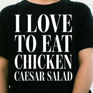 May include: A black short-sleeved t-shirt featuring the white text I LOVE TO EAT CHICKEN CAESAR SALAD printed in a bold, serif font across the chest. The design is centered and occupies the majority of the front of the garment.