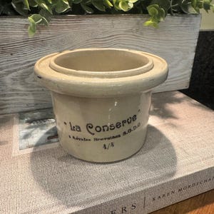 May include: A cream-coloured ceramic jar with a wide rim and a slightly flared base. The jar has the words "la Conserve" and other text printed on the side. The jar is sitting on a book.