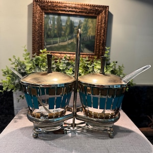 May include: A vintage salt and pepper set with a silver-tone metal frame. The glass containers have gold lids and teal and gold mosaic-style designs. Clear spoons rest inside each container. A framed landscape painting is in the background.