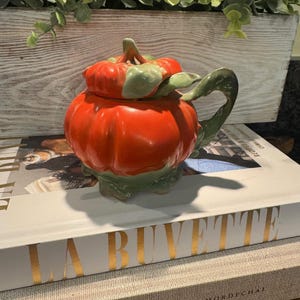 May include: A ceramic teapot shaped like a pumpkin, with a bright orange body and green accents on the lid, handle, and base. The teapot is sitting on top of a stack of books. Decorative greenery is in the background.