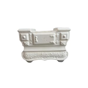 May include: A white ceramic planter shaped like a vintage wood-burning stove. The front features a central door with a handle, square side panels, and a base decorated with embossed floral and scroll patterns. The top has an open rectangular cavity.