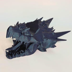 May include: A dark blue dragon skull sculpture with sharp teeth and horns. The detailed, 3D-printed fantasy art piece has a textured surface and is set against a white background. The dragon head is open-mouthed.