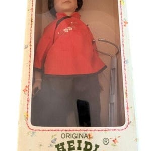 Könnte beinhalten: Eine Heidi Ott Puppe in einer Box. Die Puppe hat schwarzes Haar und trägt eine rote Jacke mit weißen Blumenakzenten und eine schwarze Hose. Auf der Box steht "Original Heidi Ott Swiss Design Handmade".