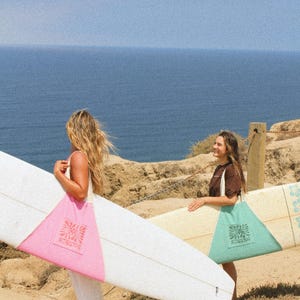 May include: Two people carrying surfboards and tote bags on a sunny day. One person holds a white surfboard with a pink accent and a pink tote bag with a logo. The other person carries a surfboard and a teal tote bag with a logo. The ocean is visible in the background.