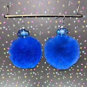 May include: A pair of vibrant blue pom-pom earrings. Each earring features a large, fluffy blue pom-pom suspended from a silver hook. The top of each earring has a cluster of blue beads and a central iridescent bead. The background is black with colorful star shapes.