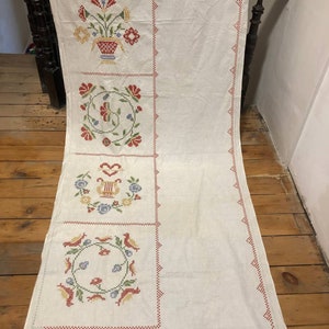 May include: A white linen tablecloth with a red, green, and blue embroidered design. The design features a vase of flowers, a circle with birds, and a circle with a lyre.