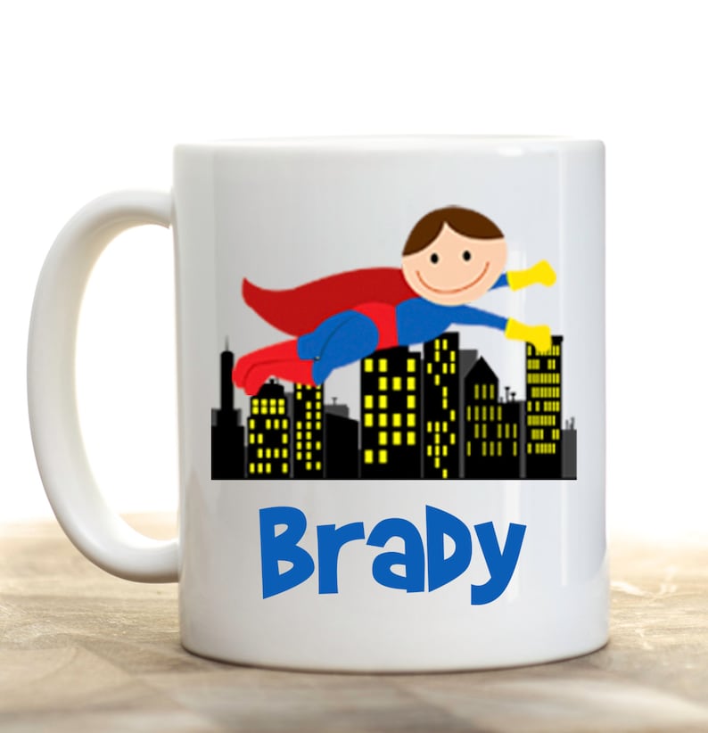 May include: White ceramic mug featuring a cartoon superhero flying over a city skyline. The superhero wears a red cape and blue suit. The name "Brady" is printed in blue, beneath the city illustration.