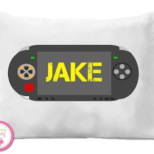 May include: White pillow with a black video game controller design. The controller has a screen with the name "JAKE" in yellow. The pillow has a pink and white logo in the bottom left corner.