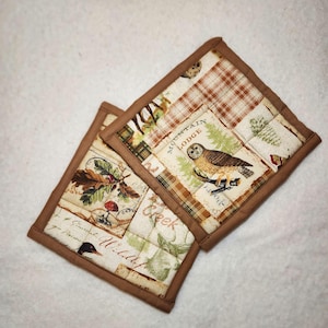 May include: Two square quilted pot holders with a brown border. The fabric features a nature-themed design with an owl, trees, and the words "Mountain Lodge" and "Creek Wildlife".
