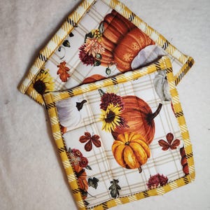 May include: Two quilted pot holders with a fall theme. The pot holders are white with a plaid pattern and feature a pumpkin, sunflower, and leaf design. The pot holders have a yellow and black trim.