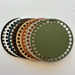 May include: A collection of seven round, flat, solid-colored bases for crochet or knitting projects. Colors include black, white, navy, brown, tan, silver, and olive green. Each base has evenly spaced holes around the perimeter.
